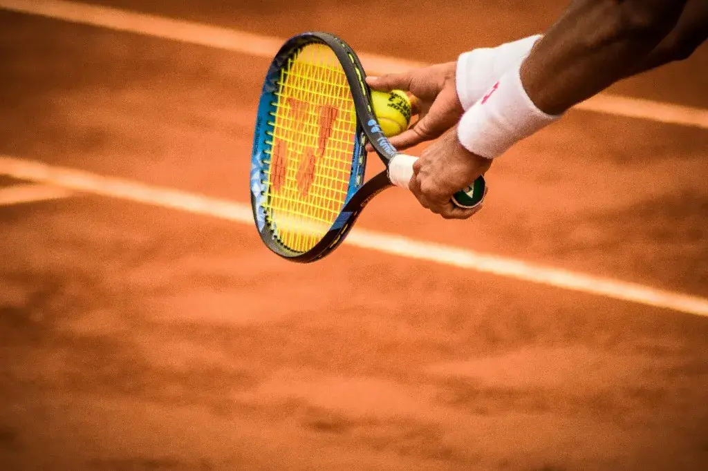 Close-up of a tennis player holding a blue and black Yonex racket on a red clay court, preparing to return a serve.
